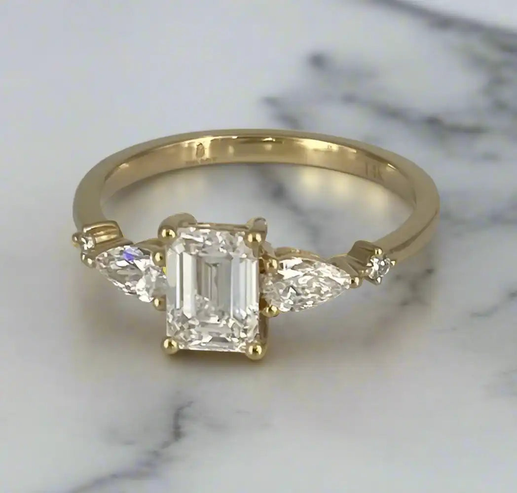 Haggith Engagement Ring with emerald-cut diamond and two side stones on 14K yellow gold band resting on a polished marble with black lightning color