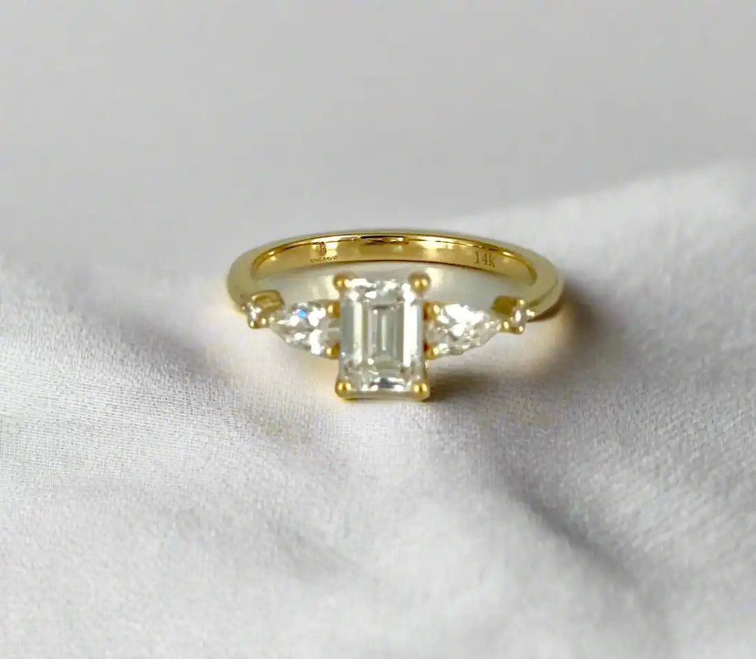 Haggith Engagement Ring with emerald-cut diamond and two side stones on 14K yellow gold band resting on soft white fabric