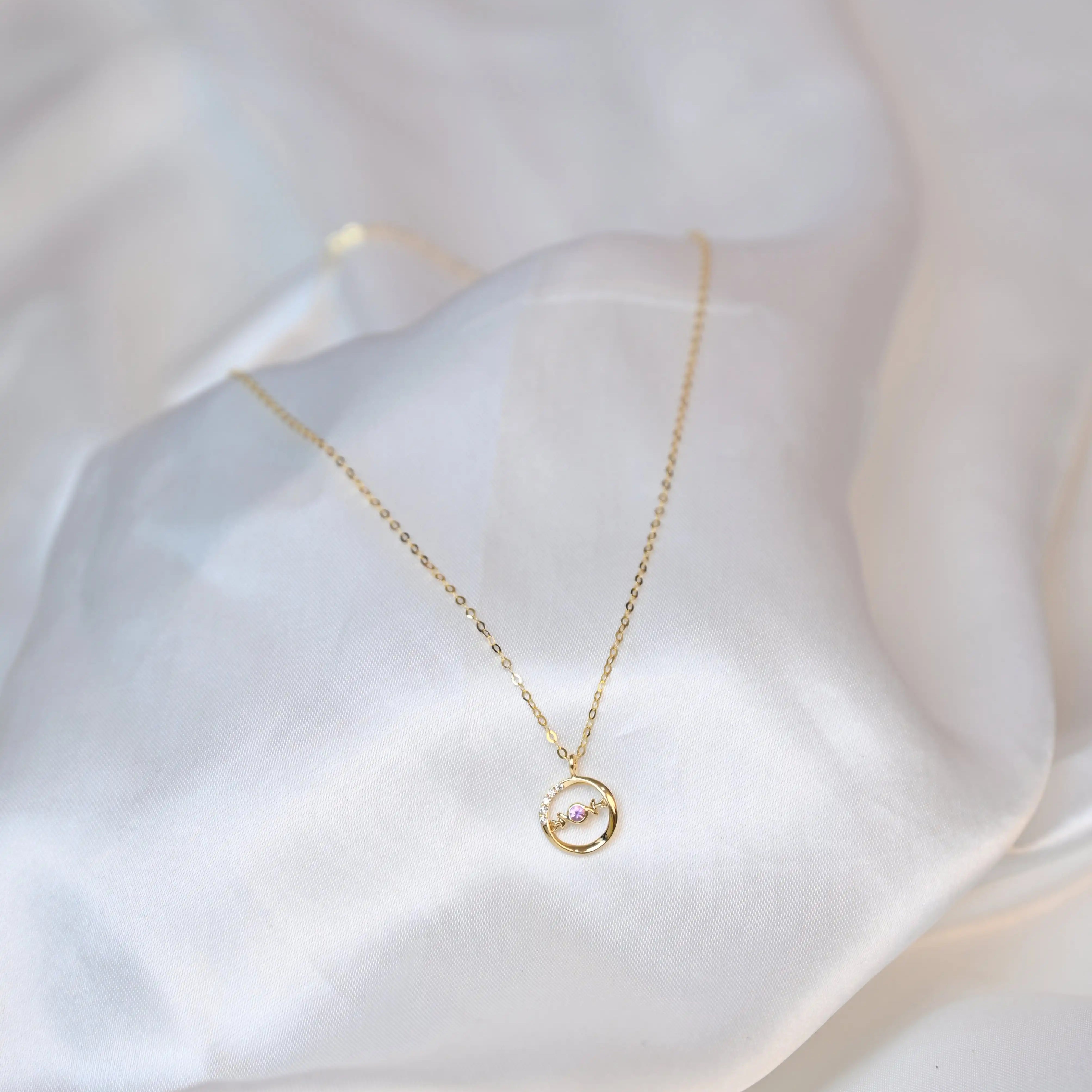 A delicate 18K yellow gold necklace with a circular pendant featuring a round pink sapphire in the center and a curved row of small white diamonds on one side. The pendant hangs from a thin gold chain on a white silk cloth