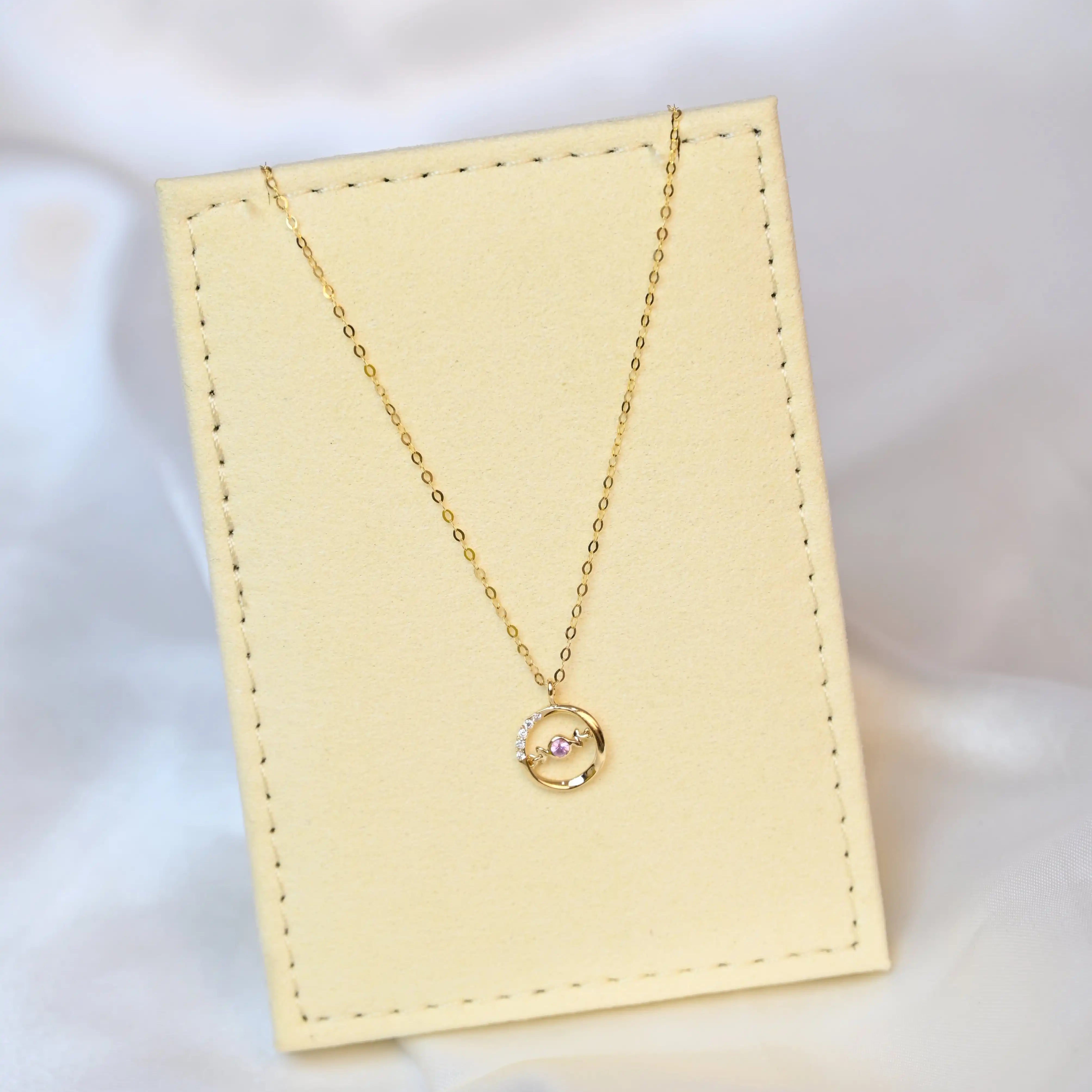A delicate 18K yellow gold necklace with a circular pendant featuring a round pink sapphire in the center and a curved row of small white diamonds on one side. The pendant hangs from a thin gold chain.