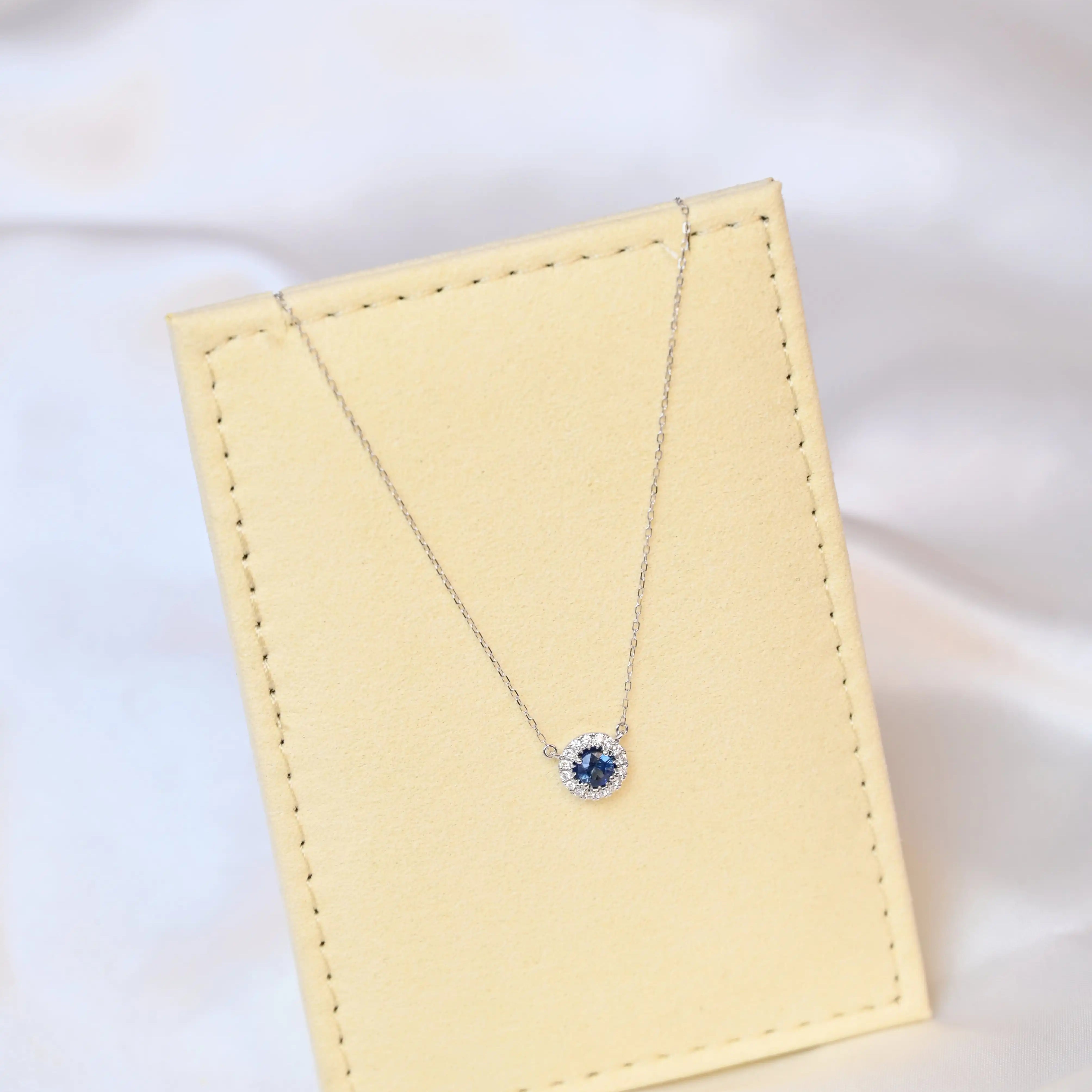 Michal Royalty Sapphire Necklace with royal blue center stone and round diamond halo in 18K white gold – luxury gemstone pendant for women on a beige jewelry display