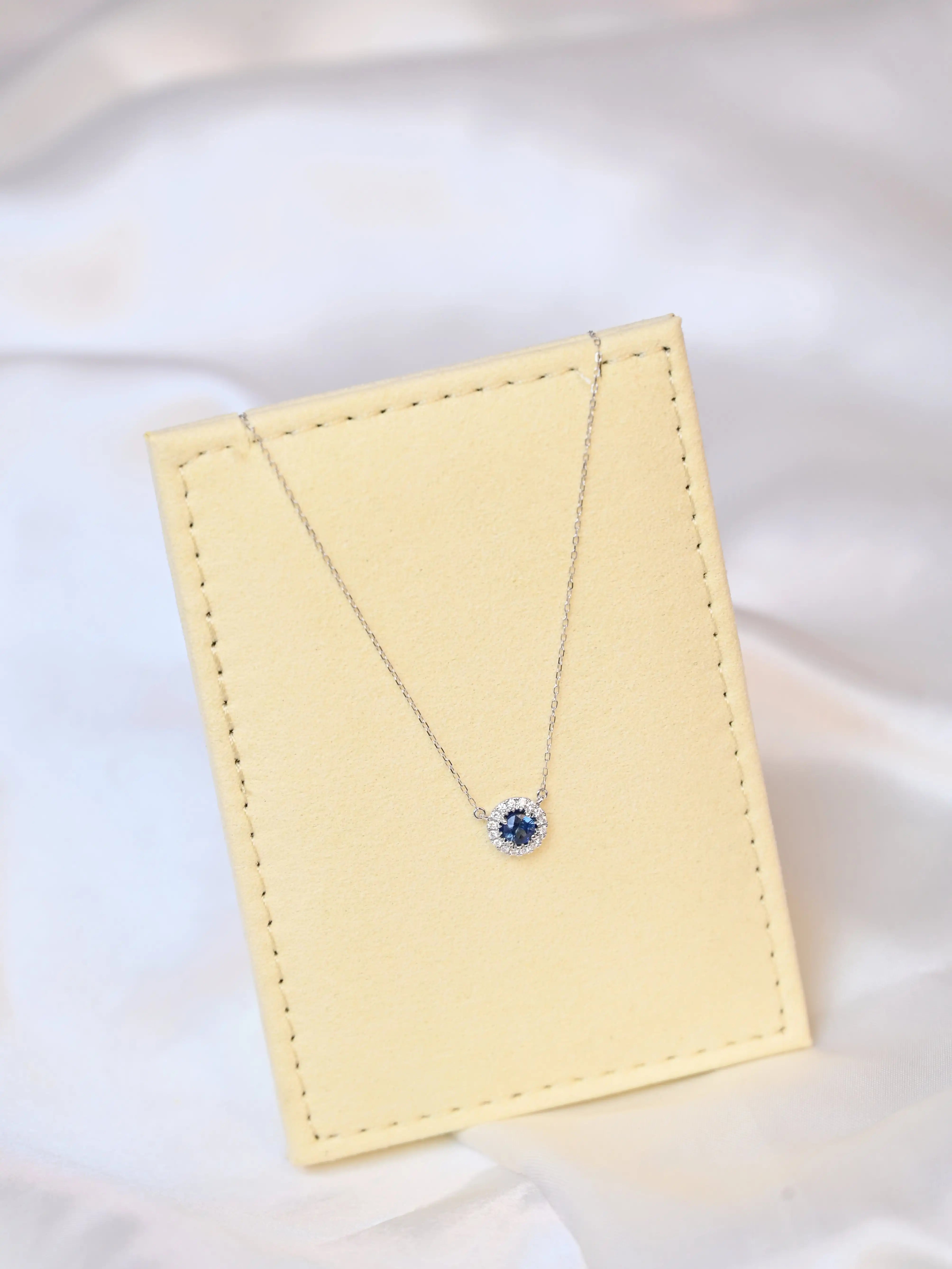 Michal Royalty Sapphire Necklace with royal blue center stone and round diamond halo in 18K white gold – luxury gemstone pendant for women on a beige jewelry display