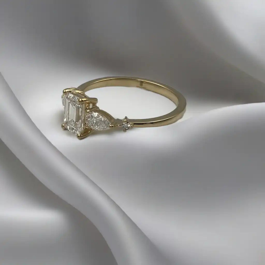 Haggith Engagement Ring with emerald-cut diamond and two side stones on 14K yellow gold band resting on soft white silk