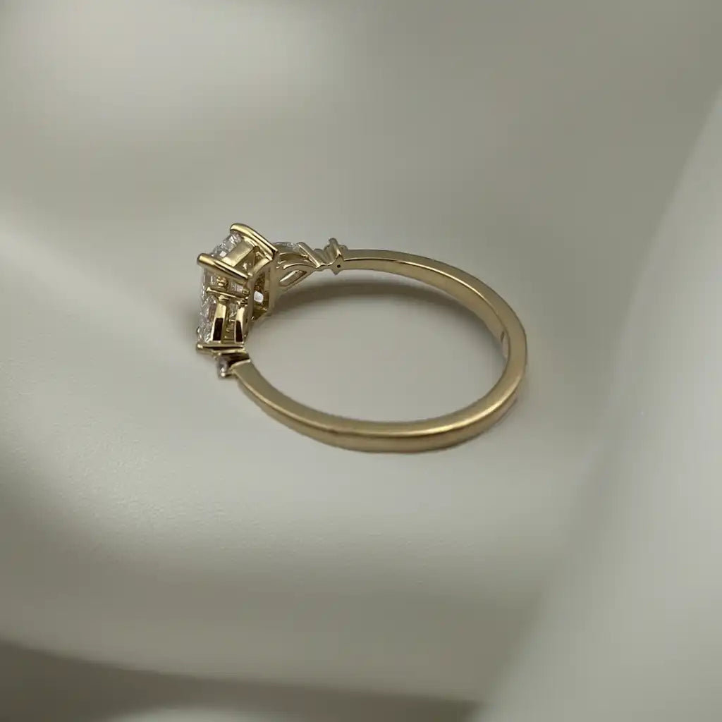 Haggith Engagement Ring with emerald-cut diamond and two side stones on 14K yellow gold band resting on soft white silk