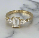Haggith Engagement Ring with emerald-cut diamond and two side stones on 14K yellow gold band resting on a polished marble with black lightning color