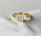 Haggith Engagement Ring with emerald-cut diamond and two side stones on 14K yellow gold band resting on soft white fabric