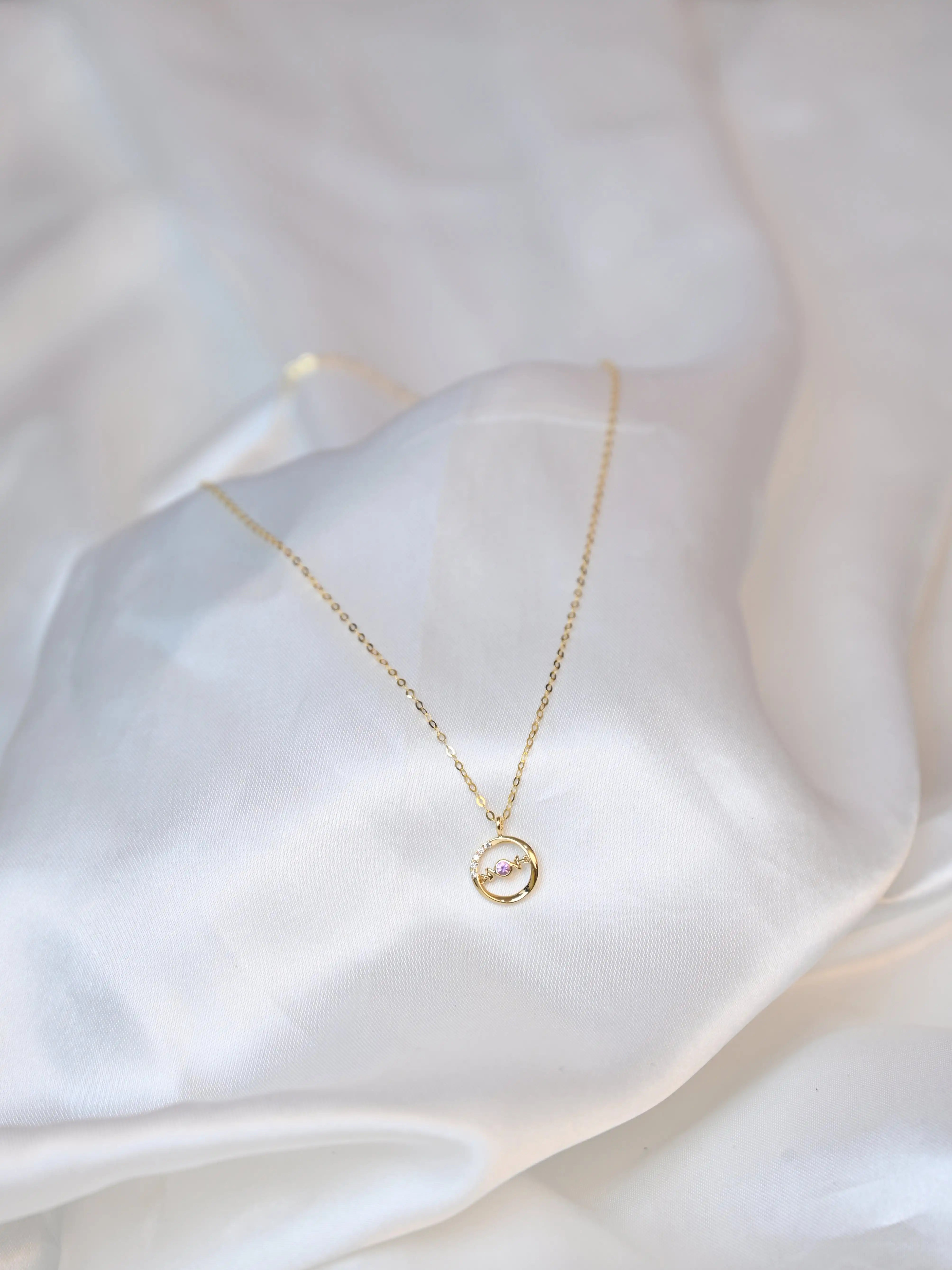 A delicate 18K yellow gold necklace with a circular pendant featuring a round pink sapphire in the center and a curved row of small white diamonds on one side. The pendant hangs from a thin gold chain on a white silk cloth