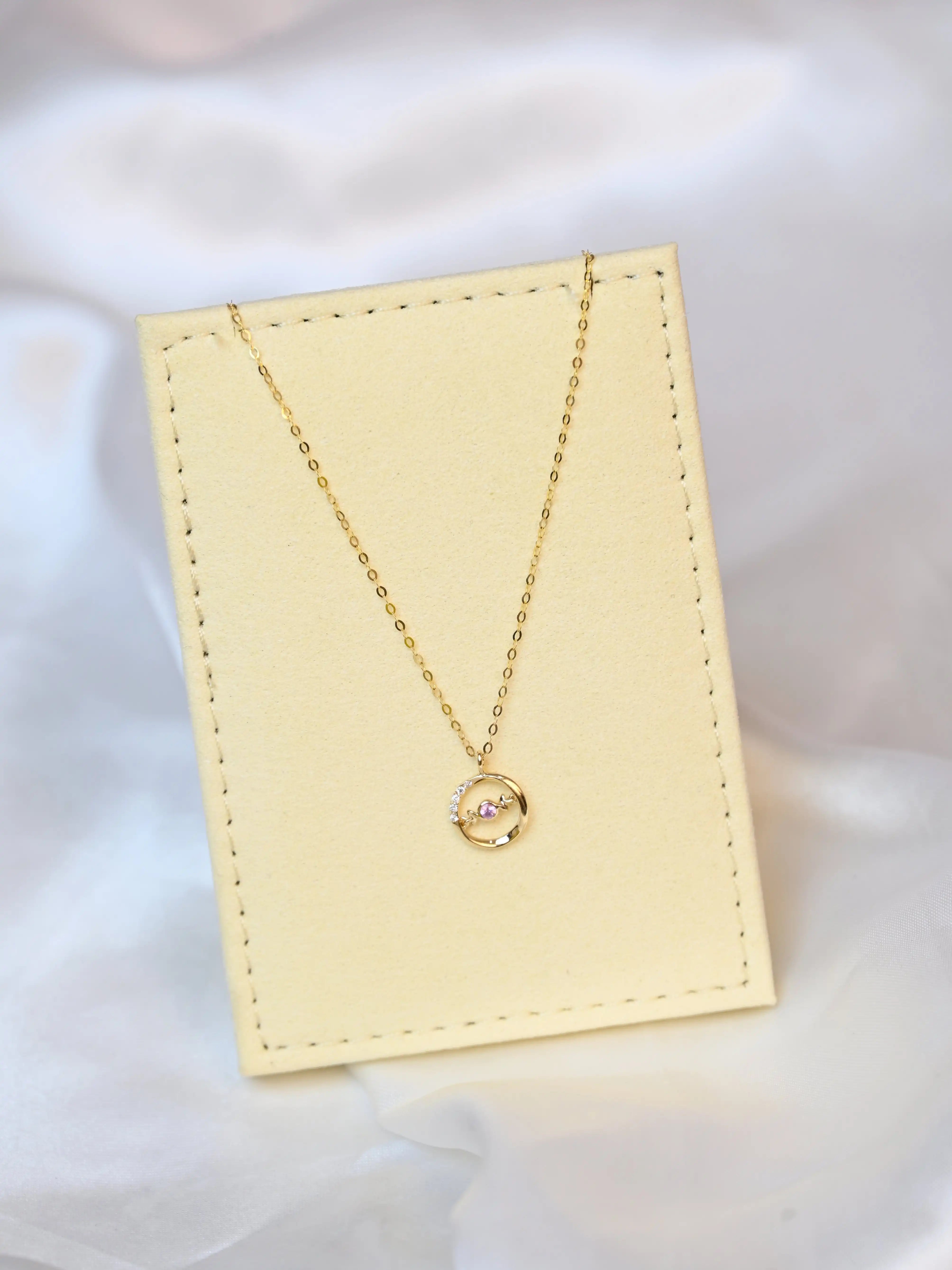 A delicate 18K yellow gold necklace with a circular pendant featuring a round pink sapphire in the center and a curved row of small white diamonds on one side. The pendant hangs from a thin gold chain.
