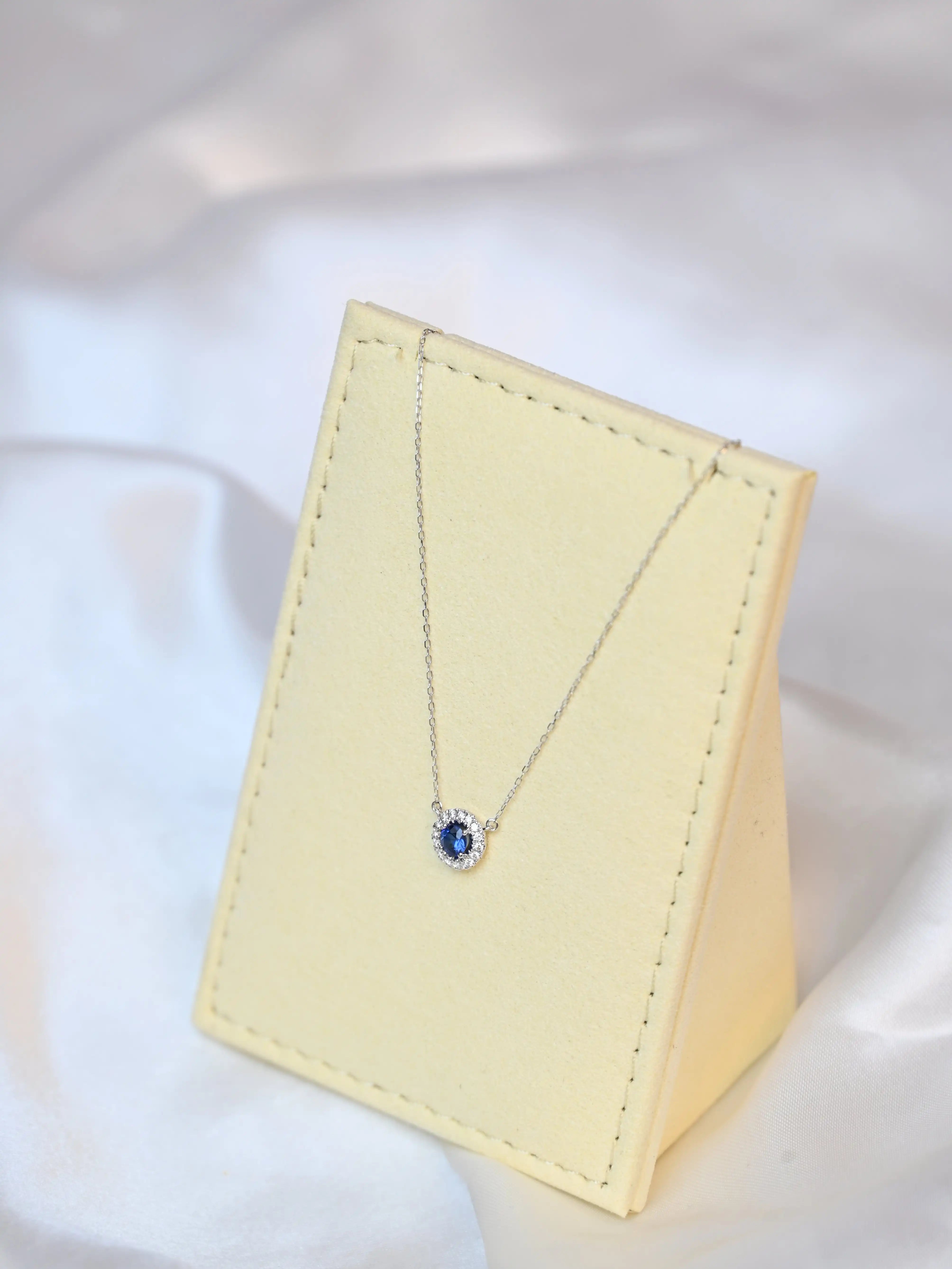 Michal Royalty Sapphire Necklace with royal blue center stone and round diamond halo in 18K white gold – luxury gemstone pendant for women on a beige jewelry display set 